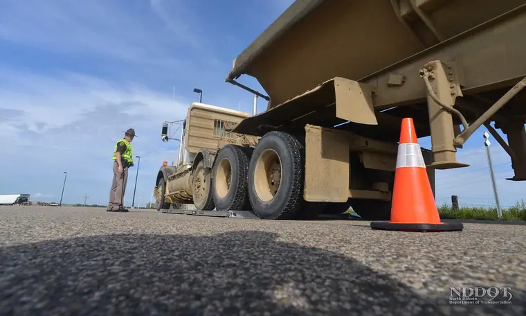 A North Dakota Highway Patrol officer beside a dump trailer stopped on portable scales, an orange cone by the wheels